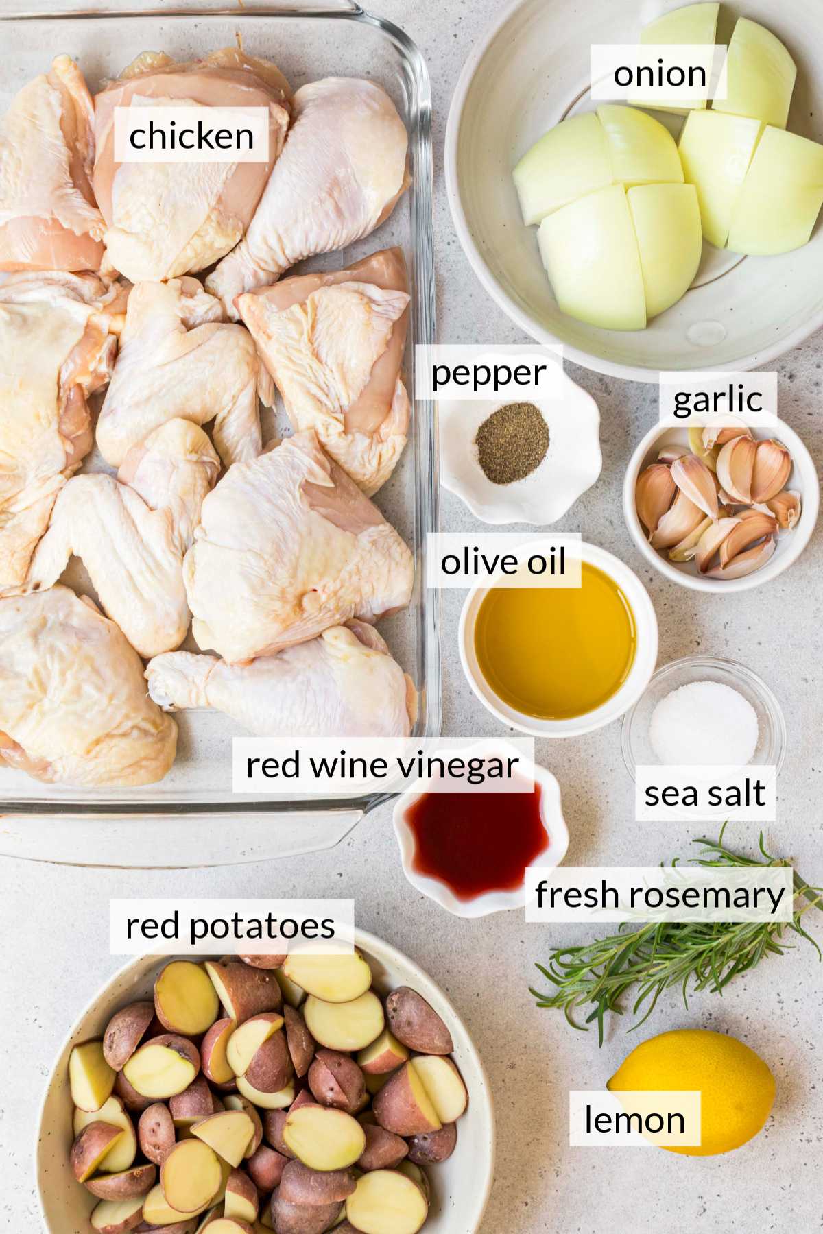 Uncooked chicken in a glass dish near small bowls of potatoes, olive oil, vinegar, garlic, onion and salt.