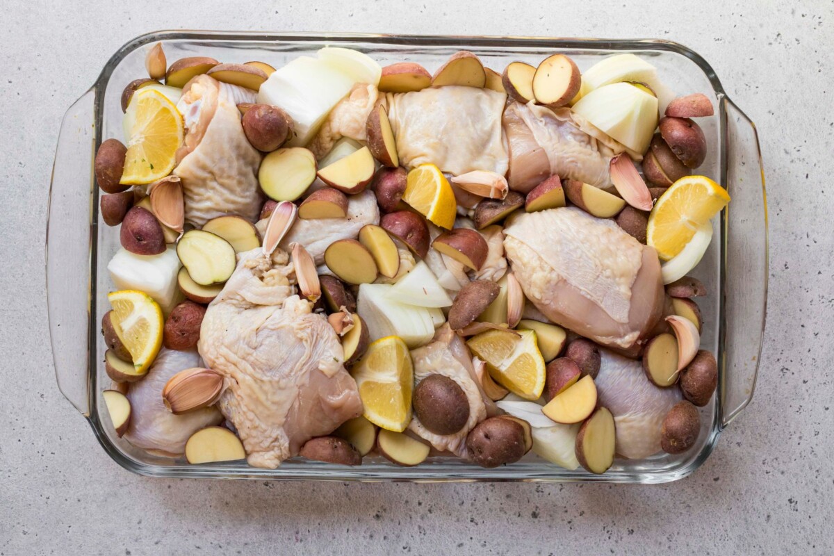 Uncooked chicken, potatoes and onion wedges in a glass baking dish.