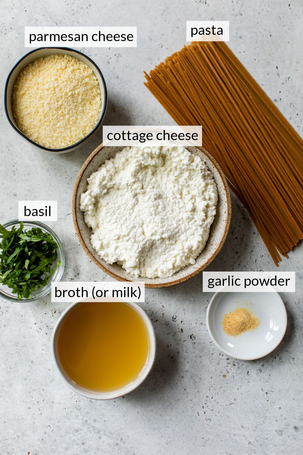 Uncooked pasta near bowls with cottage cheese, broth, parmesan cheese and basil.