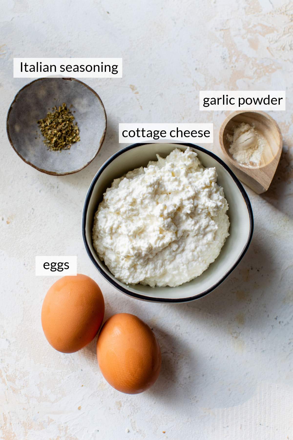 Bowl of cottage cheese near 2 eggs and small bowls of garlic powder and Italian seasoning.