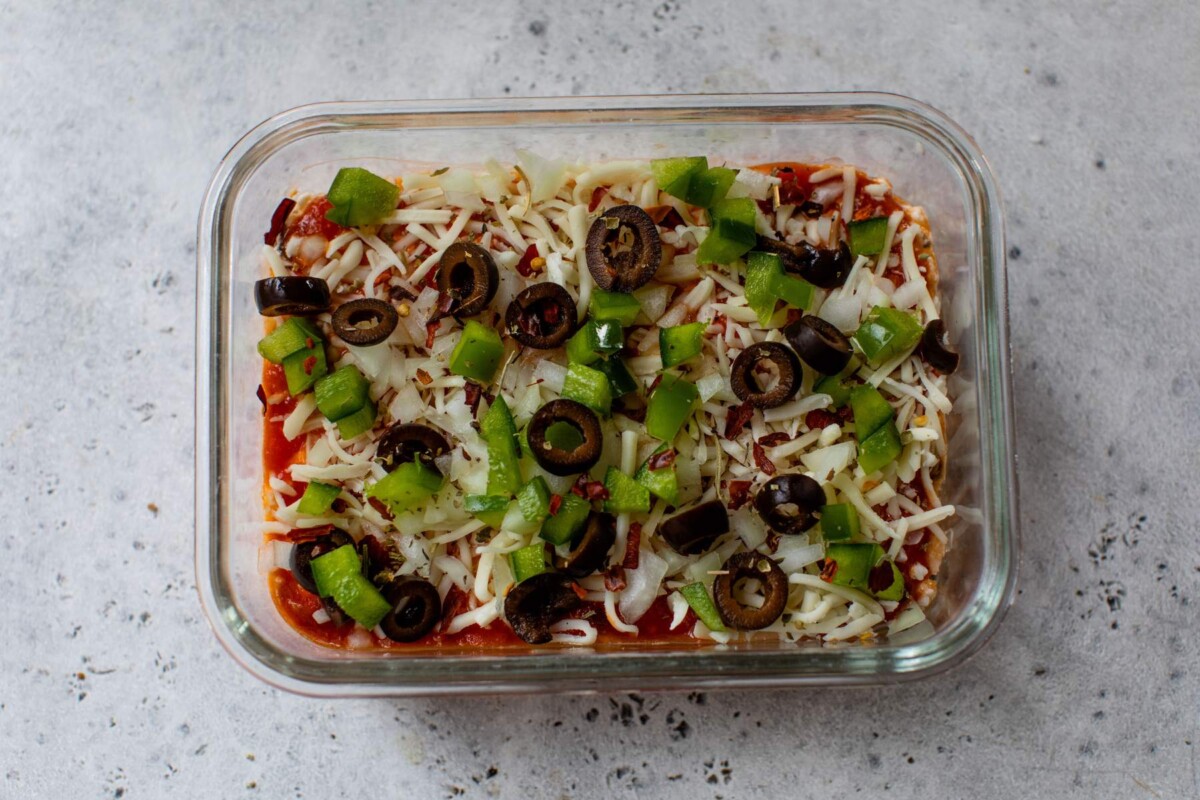 Assembling pizza bowl with black olives and chopped bell peppers.