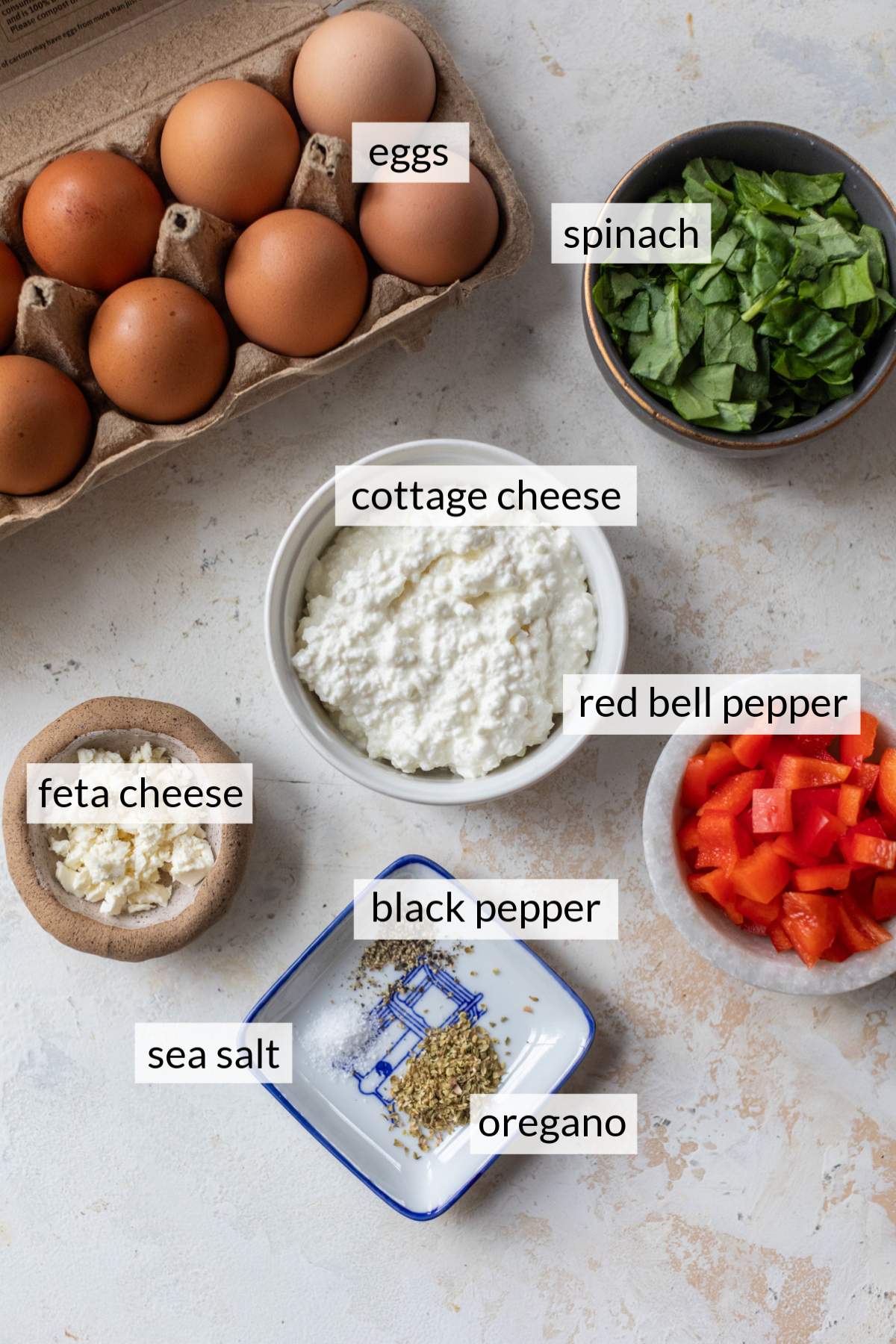 Bowls with cottage cheese, spinach, red bell pepper and spices near a carton of eggs.
