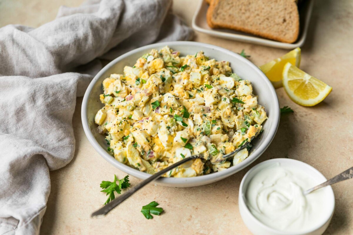 Egg salad in a white bowl near yogurt and slices of bread.