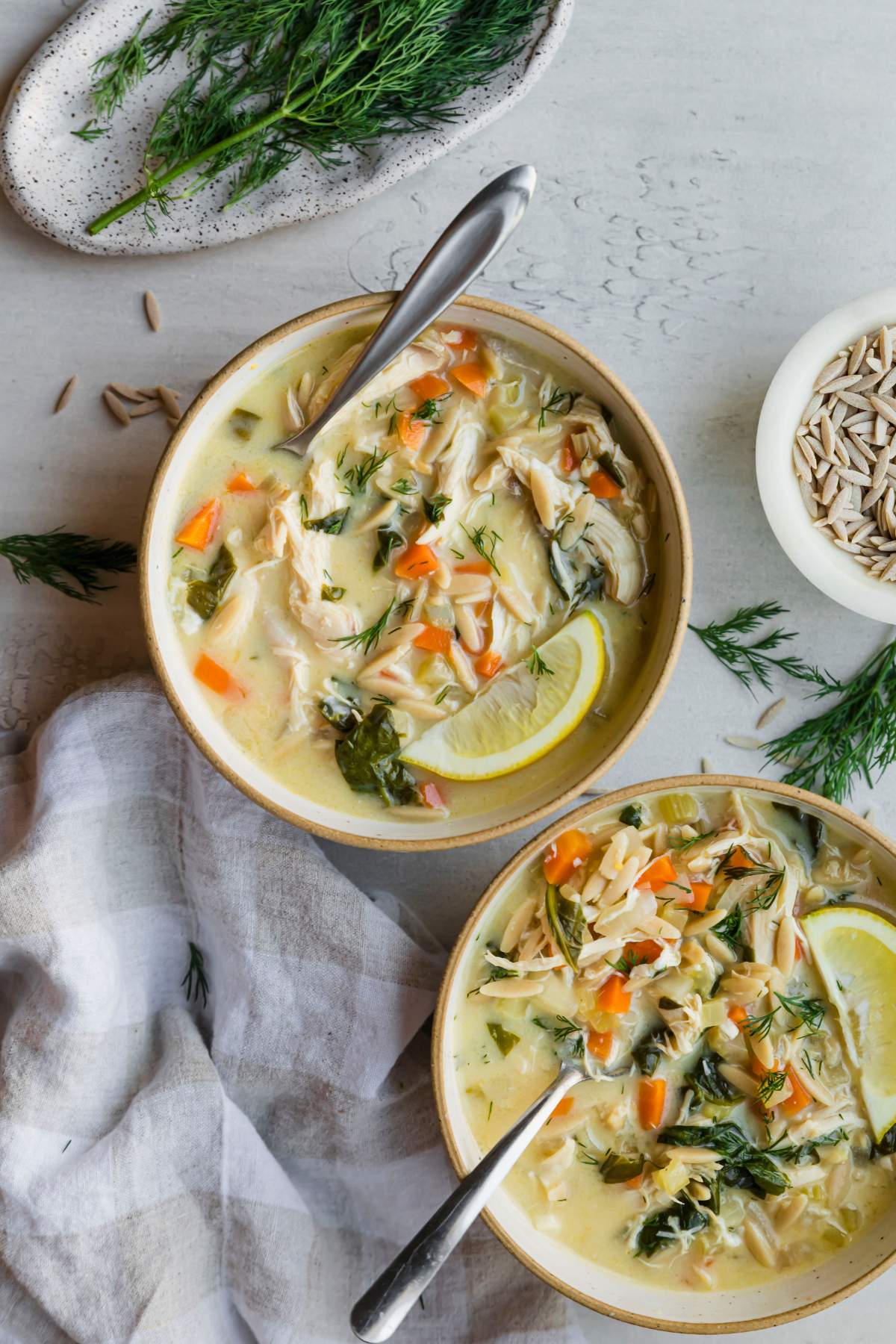 Two bowls of lemon chicken soup near fresh dill and dry orzo.