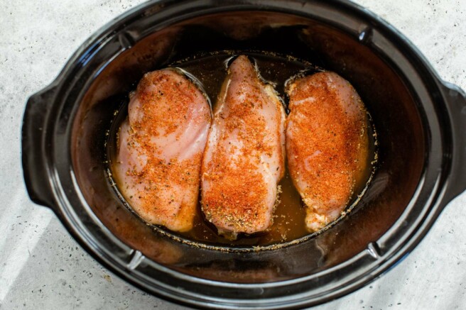 Three chicken breasts with seasoning in a slow cooker.
