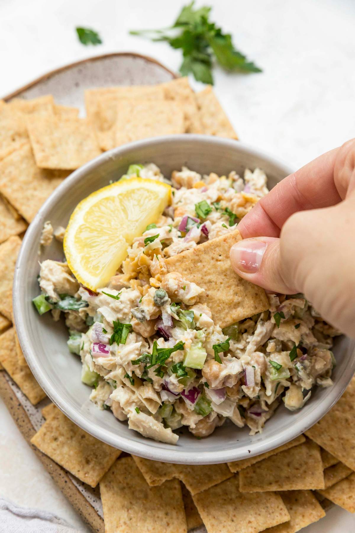 Woman's hand dipping a cracker into chickpea tuna salad in a small white bowl.